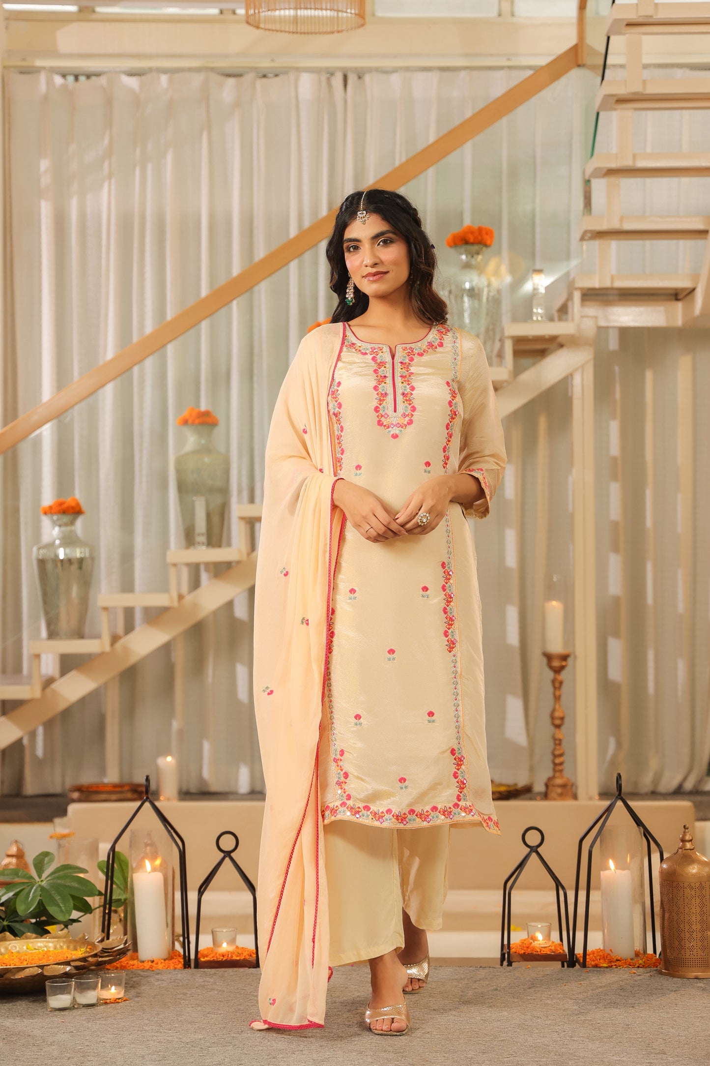 A woman in the Nivishas - Sec46 Elegant Glow Beige Embroidered Kurta Set with Dupatta stands indoors near a staircase, surrounded by lit candles and floral decor, smiling gently with her hands clasped.