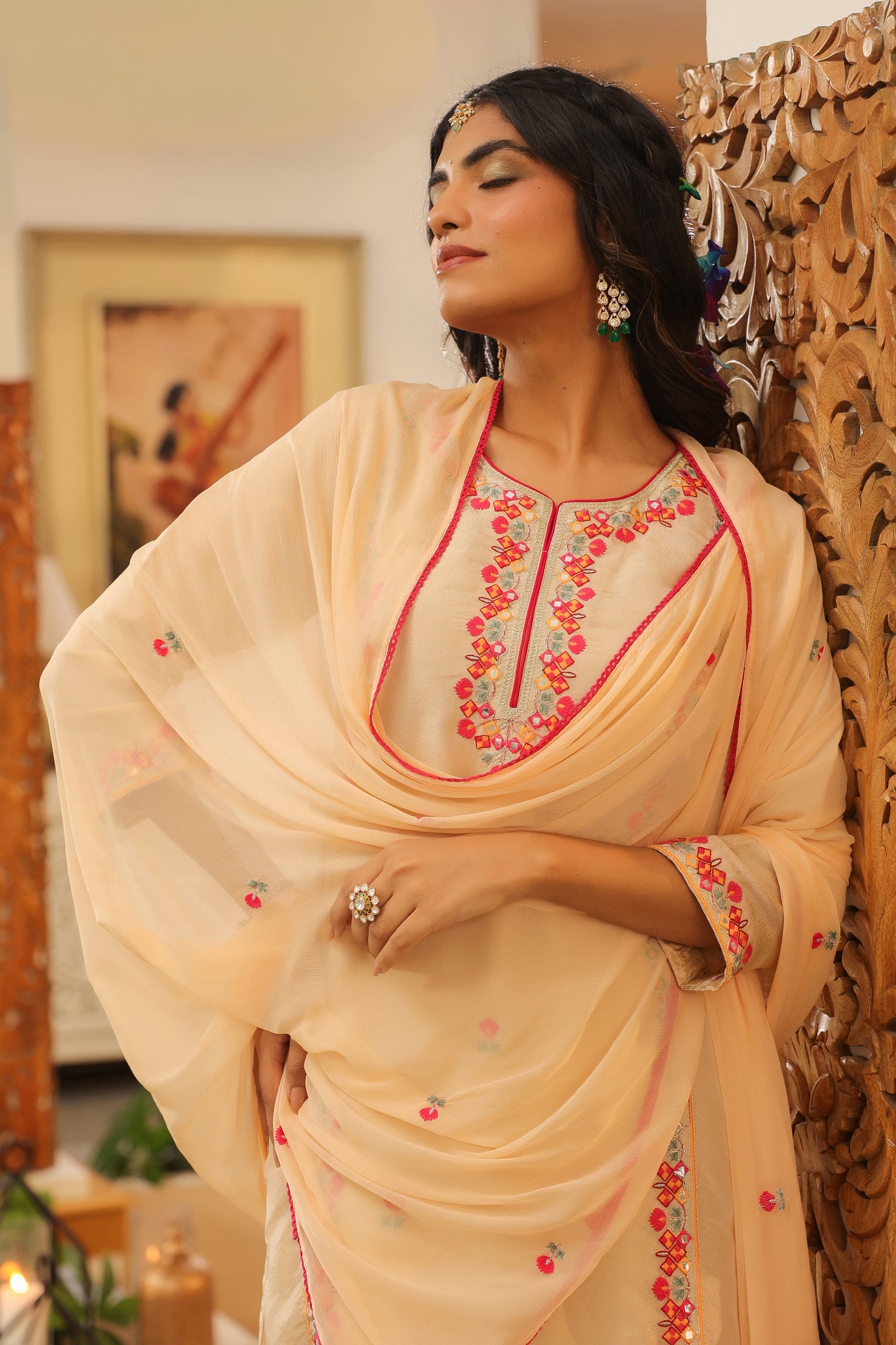 A woman poses gracefully in the Elegant Glow – Beige Embroidered Kurta Set with Dupatta by Nivishas - Sec46, featuring pink floral embroidery and styled with statement jewelry beside a carved wooden panel indoors.