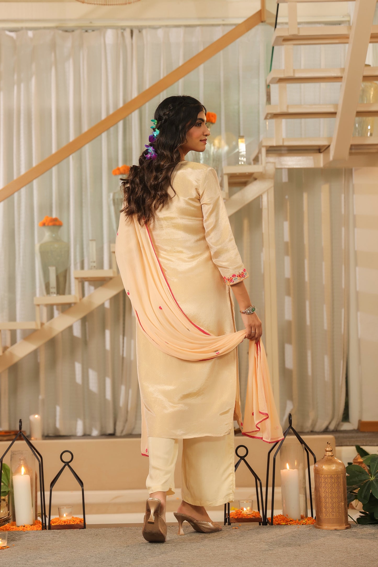 A woman wears the Elegant Glow – Beige Embroidered Kurta Set with Dupatta from Nivishas - Sec46, paired with floral hair accessories, surrounded by candles, lanterns, and marigolds near a modern staircase.