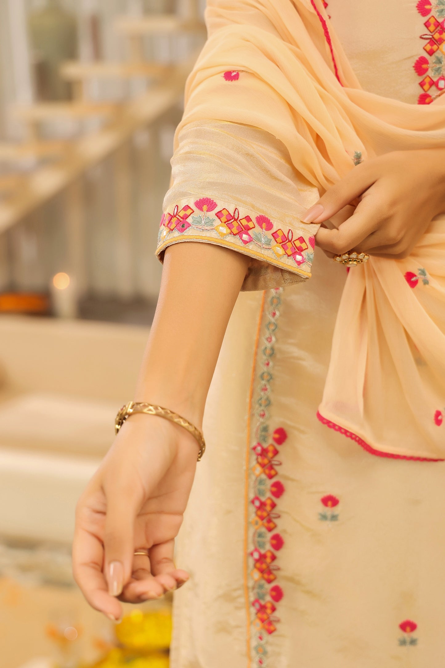 A person wears the Elegant Glow – Beige Embroidered Kurta Set with Dupatta by Nivishas - Sec46, featuring pink and yellow floral embroidery and a gold bracelet, for a graceful look in soft silk paired with a matching chiffon dupatta.