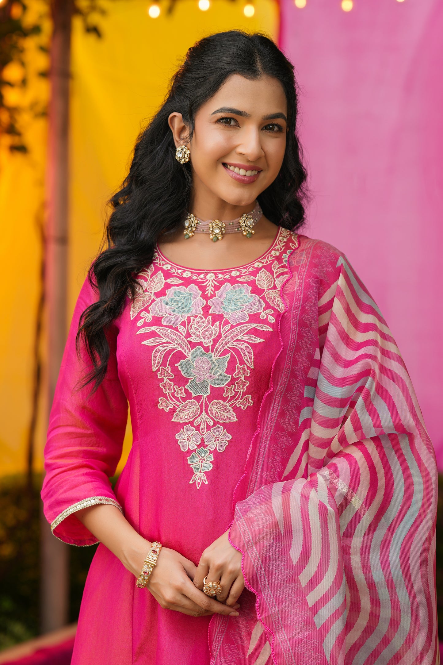 A smiling woman with long, wavy black hair wears Nivishas - Sec46's Gulabi Grace, a hot pink embroidered kurta set with a matching striped dupatta, paired with gold jewelry, against a festive yellow and pink backdrop.
