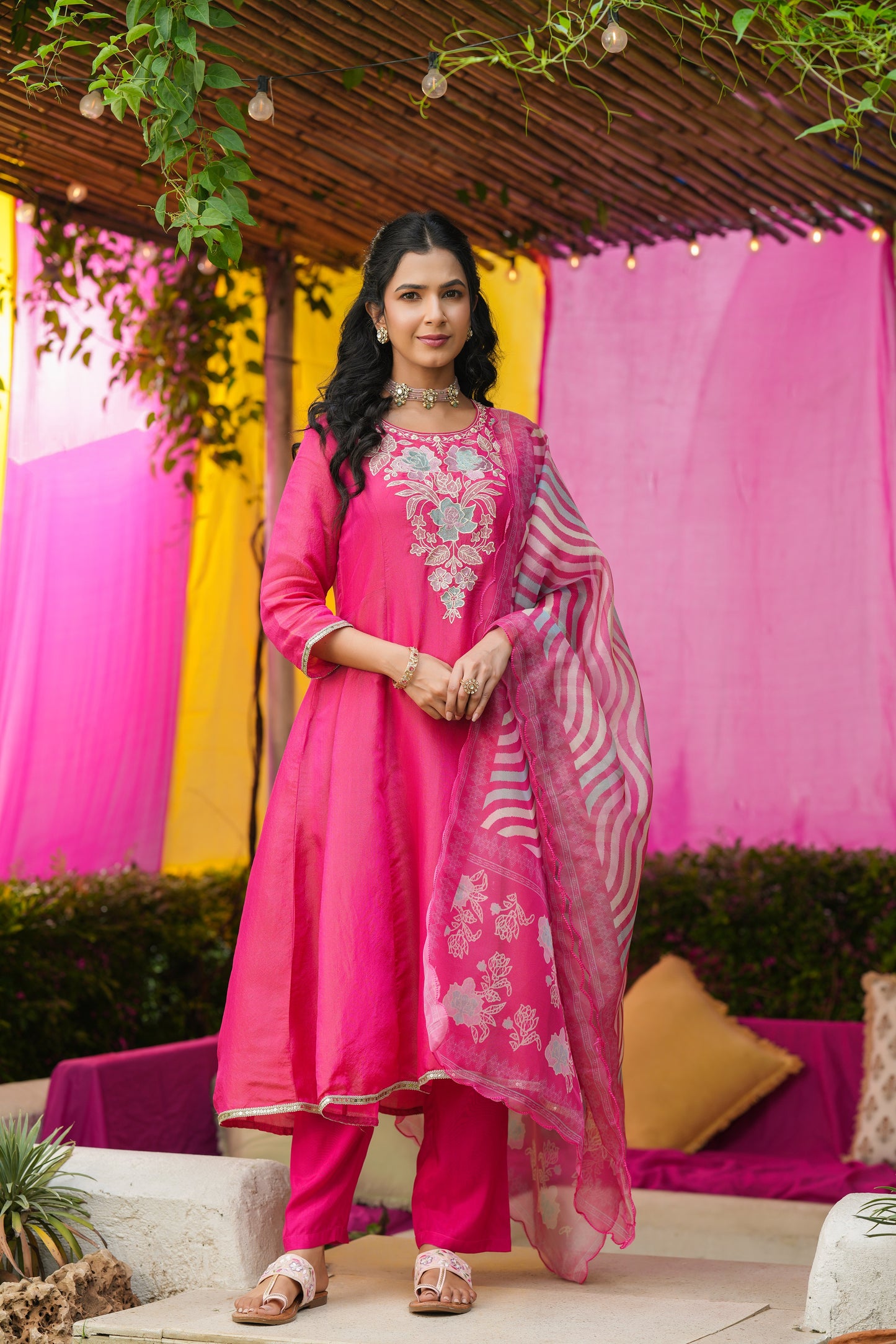 A woman stands on a patio in the Gulabi Grace – Hot Pink Embroidered Kurta Set with Striped Dupatta by Nivishas - Sec46, smiling amid pink and yellow decor and lush greenery.