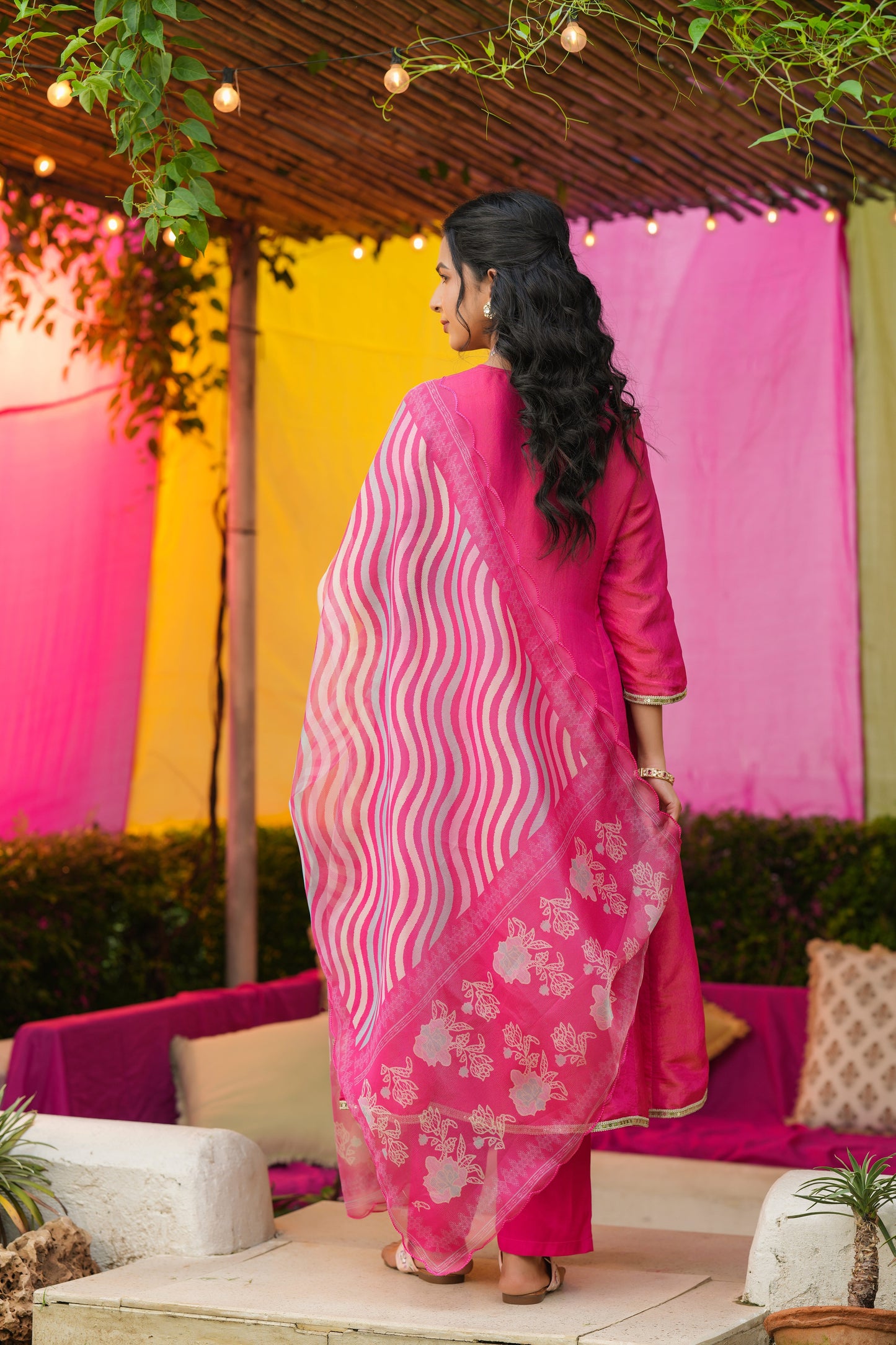A woman with long black hair, wearing the Gulabi Grace – Hot Pink Embroidered Kurta Set with Striped Dupatta from Nivishas - Sec46, stands facing away in a vibrant outdoor setting decorated with string lights.