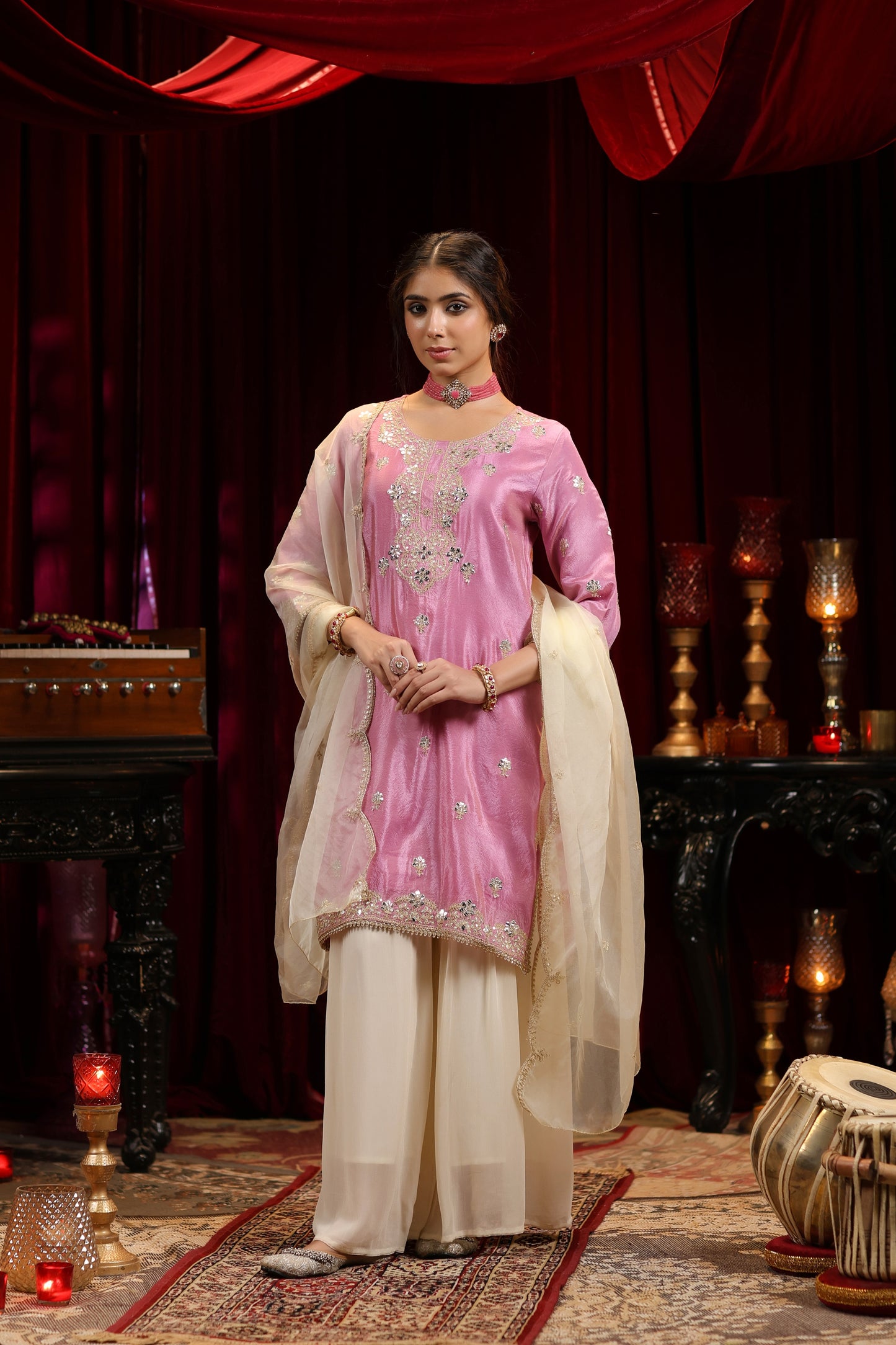 A woman wears the Nivishas - Sec46 Onion Bloom pastel pink embroidered kurta with ivory sharara and dupatta, posing indoors amid red drapes, candles, musical instruments, and ornate furniture.