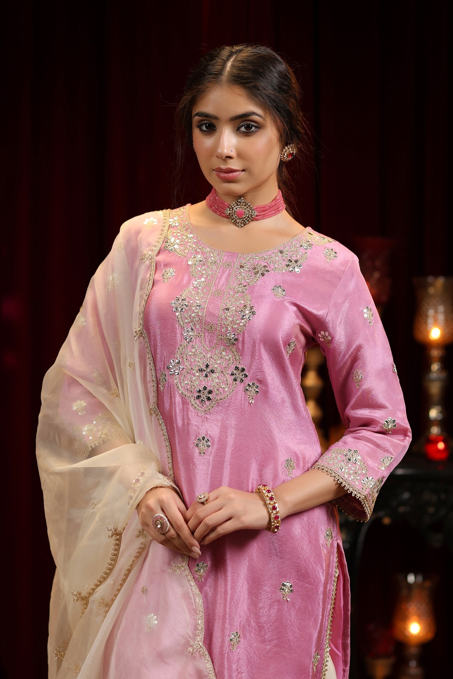 A woman poses indoors in the Onion Bloom – Pastel Pink Embroidered Kurta with Ivory Sharara & Dupatta by Nivishas - Sec46, styled with matching jewelry. The setting includes dark curtains and decorative lights.