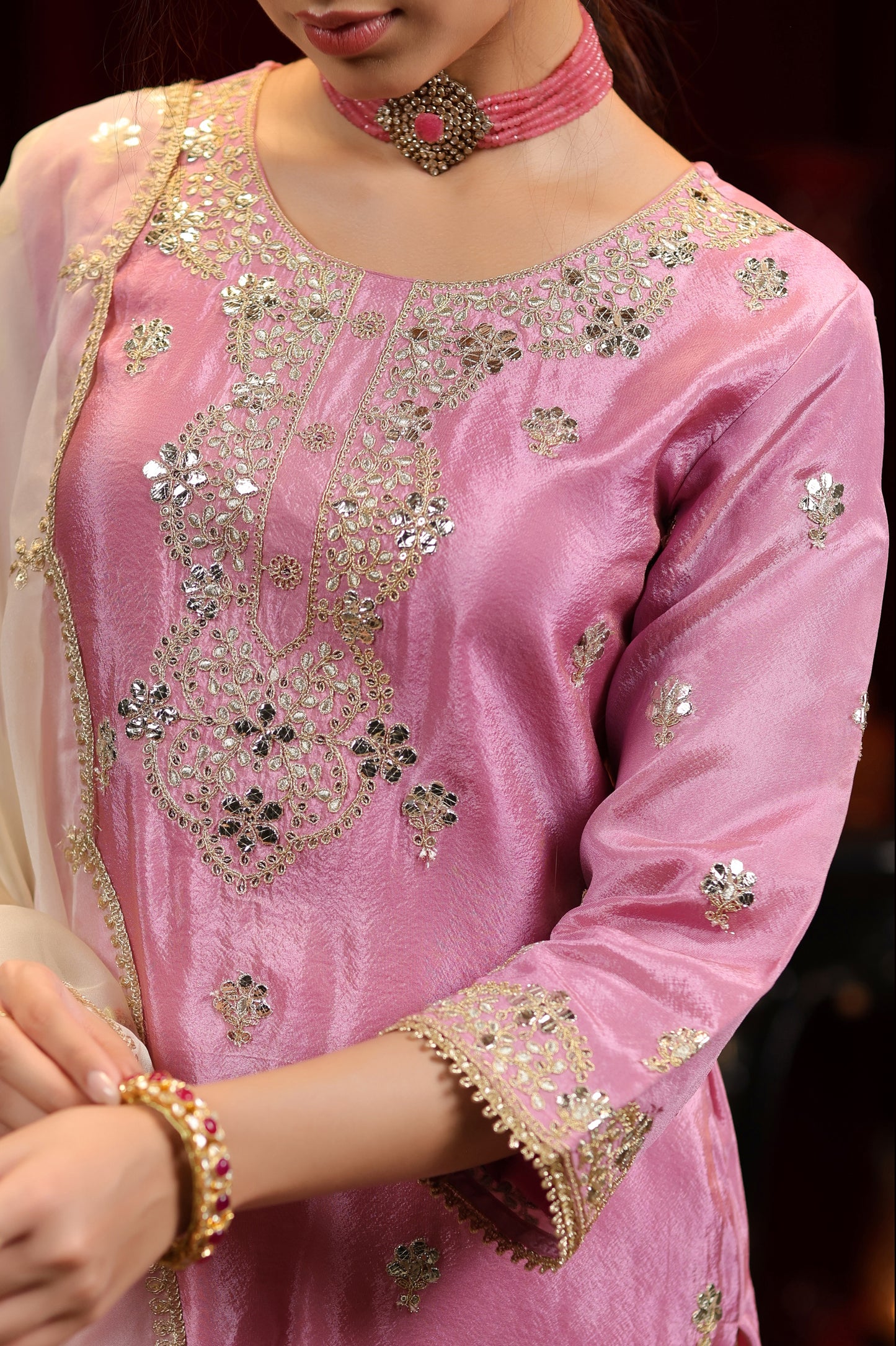 A woman models the Onion Bloom – Pastel Pink Embroidered Kurta with Ivory Sharara & Dupatta from Nivishas - Sec46, accessorized with a choker and bracelet. Her face is partially visible, turned down, shown from the shoulders down.