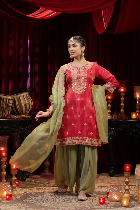 A woman dressed in Nivishas - Sec46's Scarlet Radiance, a red embroidered kurta with olive green sharara and dupatta, adorned with jewelry, stands gracefully amid candles and traditional decor.