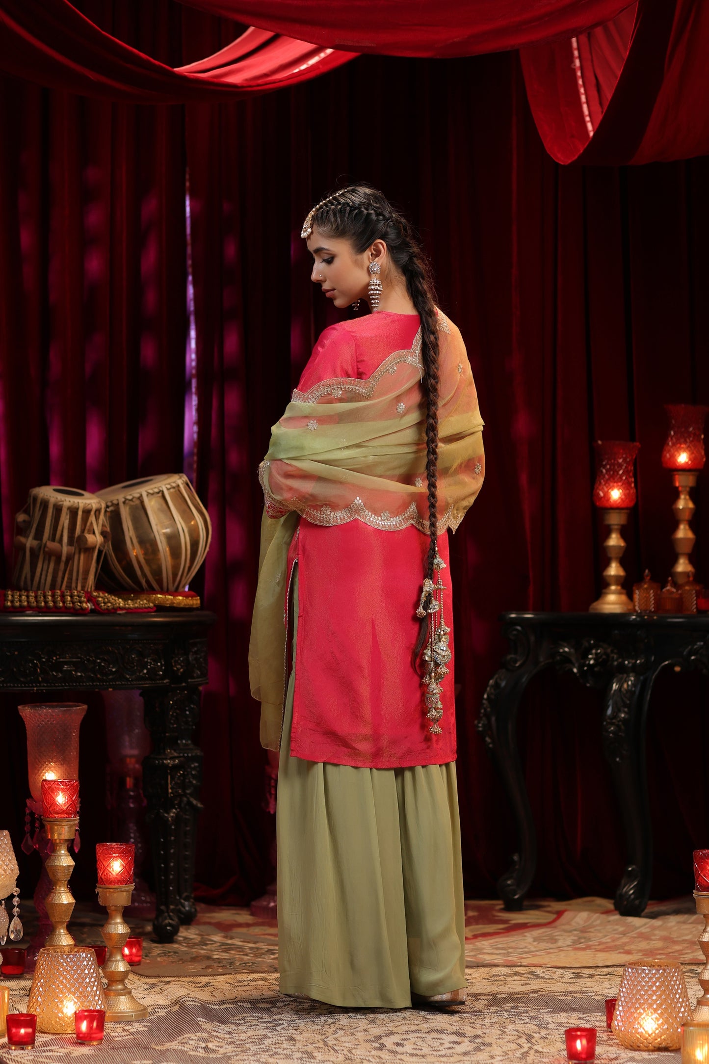 A woman in Nivishas - Sec46's Scarlet Radiance, a red embroidered kurta with olive green sharara and dupatta, stands gracefully in an opulent room adorned with red drapes, candles, and drums, her long braid cascading as she faces away.