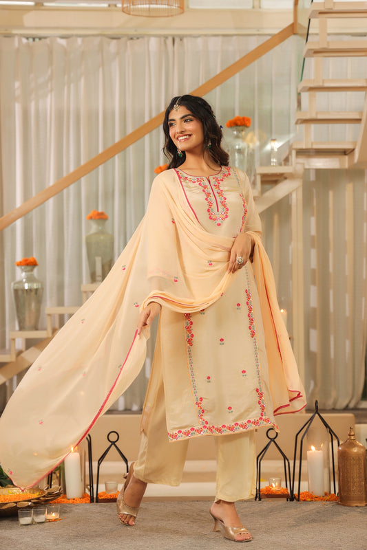 A woman smiles indoors wearing Nivishas - Sec46's Elegant Glow – Beige Embroidered Kurta Set with Dupatta, styled with sandals and jewelry. Candles and marigold flowers are arranged around her for a festive touch.