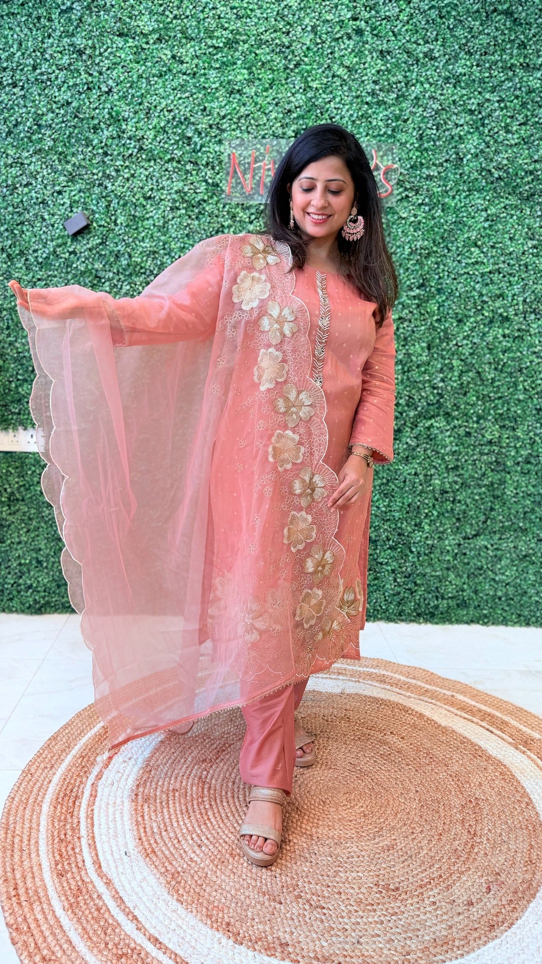 Woman in a peach traditional outfit with floral embroidery standing on a woven mat against a green textured wall.