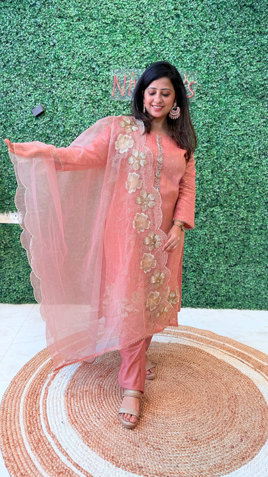 Woman in a peach traditional outfit with floral embroidery standing on a woven mat against a green textured wall.