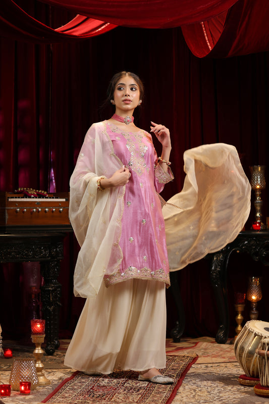 A woman showcases the Onion Bloom – Pastel Pink Embroidered Kurta with Ivory Sharara & Dupatta by Nivishas - Sec46, posing indoors amid rich red drapes and ornate decor, creating a festive and elegant atmosphere.