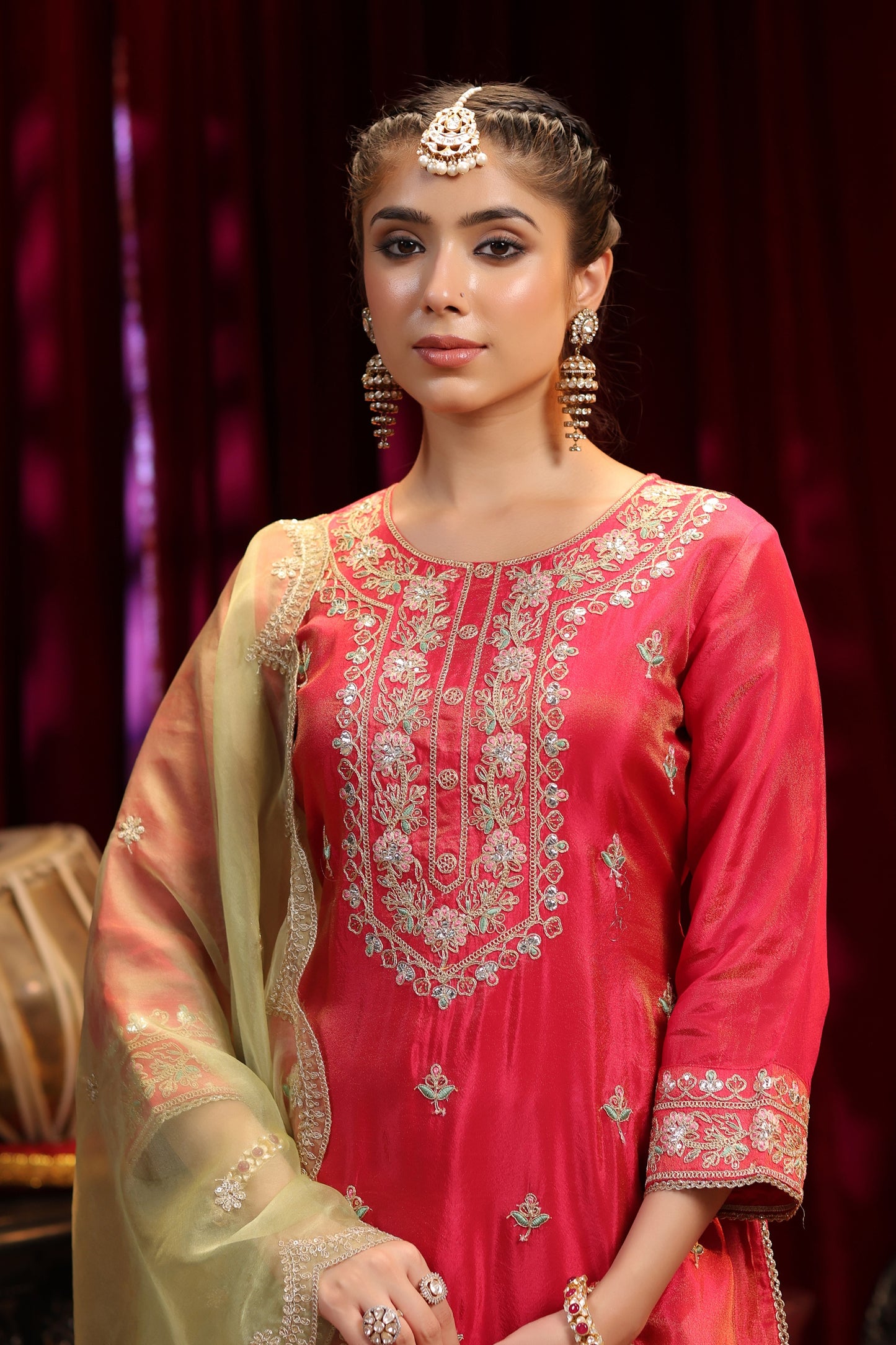A woman in Nivishas - Sec46’s Scarlet Radiance—a red embroidered kurta with olive green sharara and dupatta—stands against a deep red curtain, creating a striking festive look.