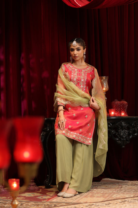 A woman glows in celebration, wearing Nivishas - Sec46's Scarlet Radiance: a red embroidered kurta with olive green sharara and dupatta, standing by lit candles and ornate decor against rich red curtains.