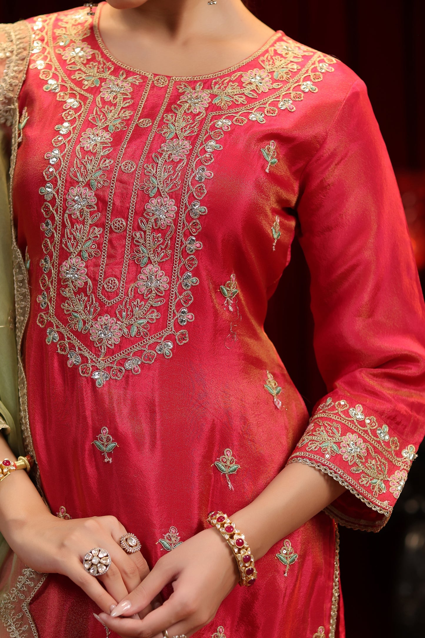 A woman in Nivishas - Sec46's Scarlet Radiance: a red embroidered kurta with olive green sharara and dupatta, featuring gold details and festive jewelry. The image focuses on her upper body and hands in this elegant ensemble.